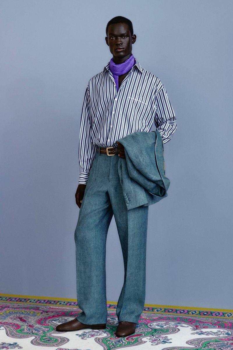Dark-skinned male model stands in a light blue suit with striped shirt and purple scarf, posing in a fashion studio setting.