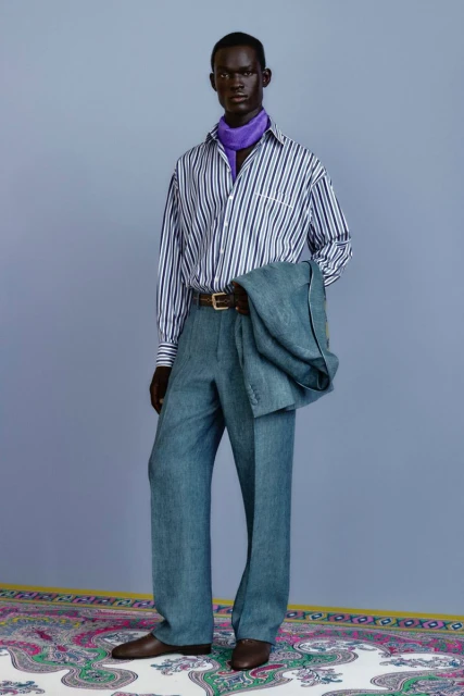 Dark-skinned male model stands in a light blue suit with striped shirt and purple scarf, posing in a fashion studio setting.