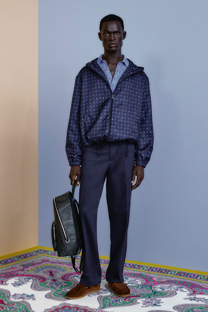 Full-length studio shot: a dark-skinned adult male wearing a navy patterned jacket, navy trousers, and carrying a backpack.
