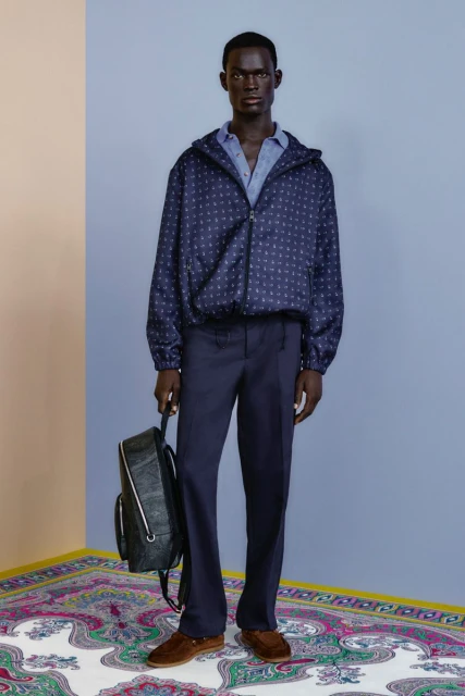Full-length studio shot: a dark-skinned adult male wearing a navy patterned jacket, navy trousers, and carrying a backpack.