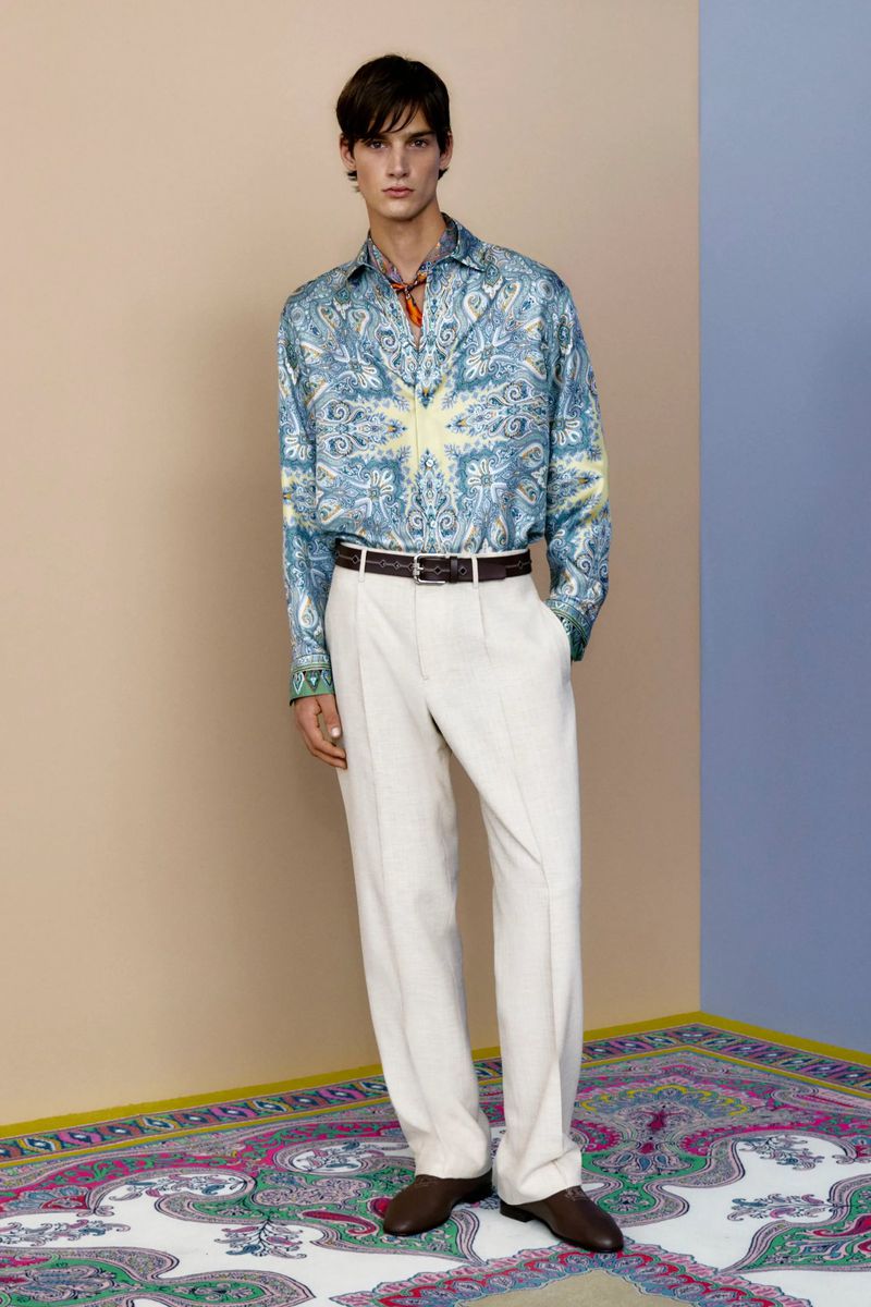 Male fashion model stands in a studio wearing a patterned button-down shirt, white pants, and brown leather dress shoes on a patterned floor.