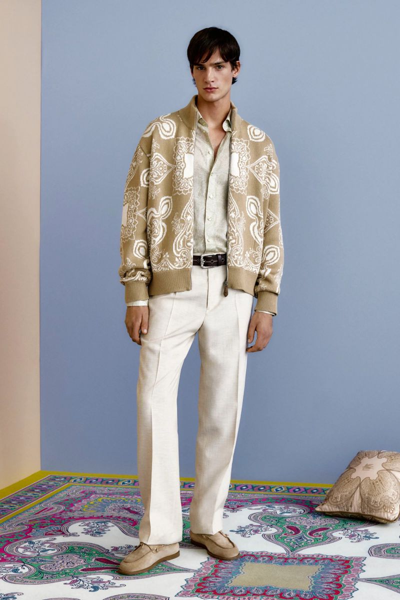 Full-length shot of a man in a paisley jacket and white trousers standing on a paisley rug against a light blue backdrop.