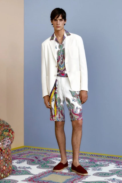 A male fashion model showcases a coordinated white jacket, patterned shirt, printed shorts, and casual loafers on a neutral backdrop.
