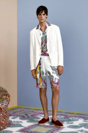 A male fashion model showcases a coordinated white jacket, patterned shirt, printed shorts, and casual loafers on a neutral backdrop.