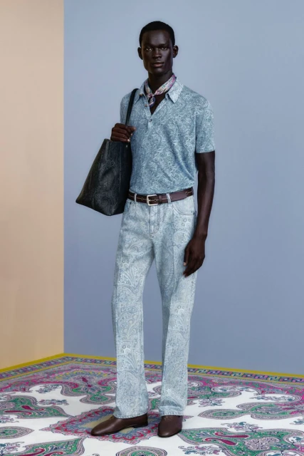 Dark-skinned male fashion model poses indoors in a patterned polo shirt, paisley pants, brown belt and shoes, holding a large tote.