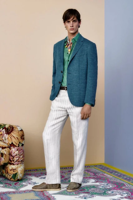 Full-length shot of a man in a turquoise blazer, green shirt, white pants, and brown shoes posing indoors on a patterned rug with a floral sofa.