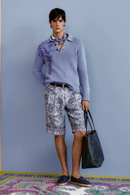 A male model poses in a blue knit sweater and paisley shorts, holding a tote bag on a patterned rug against a solid blue wall.