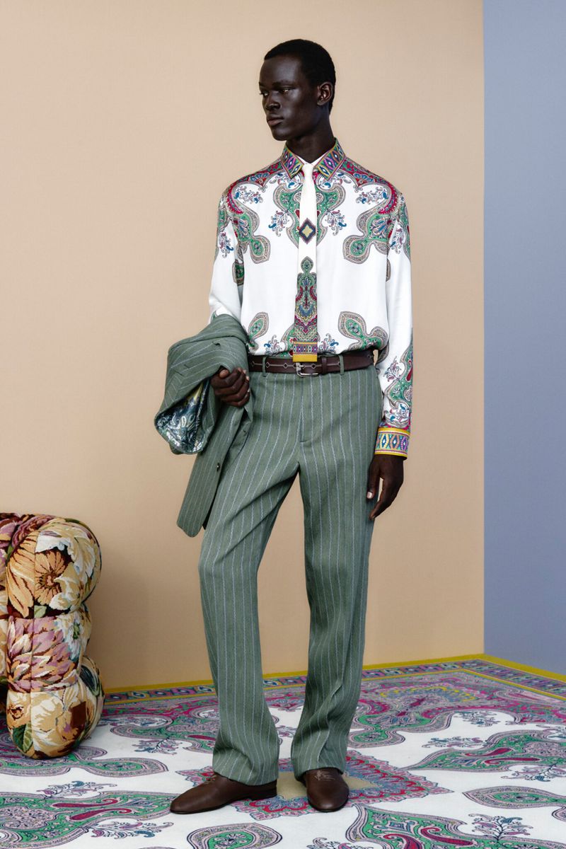 A dark-skinned male model poses indoors wearing a patterned dress shirt, pinstripe pants, brown leather shoes, and holding a jacket.