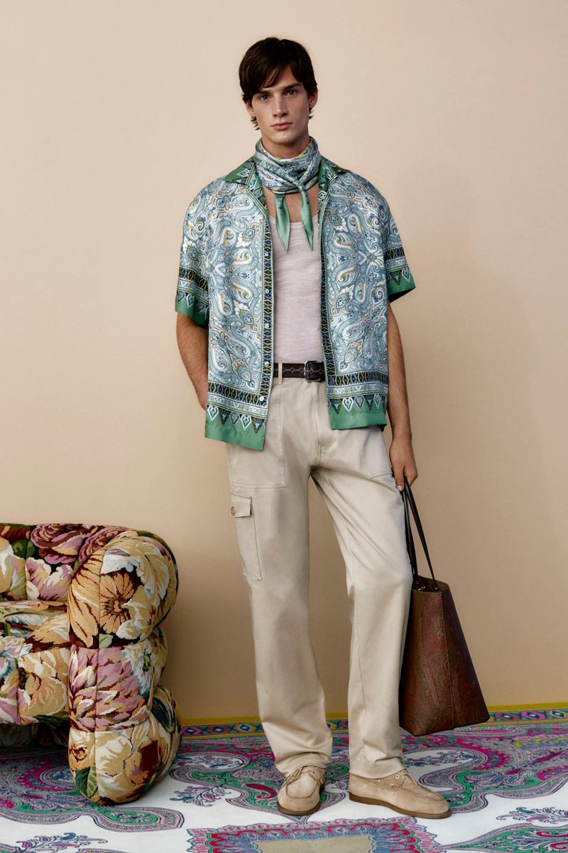 A stylish male fashion model poses in a patterned shirt, beige trousers, and loafers indoors, carrying a leather tote bag in a studio setting.