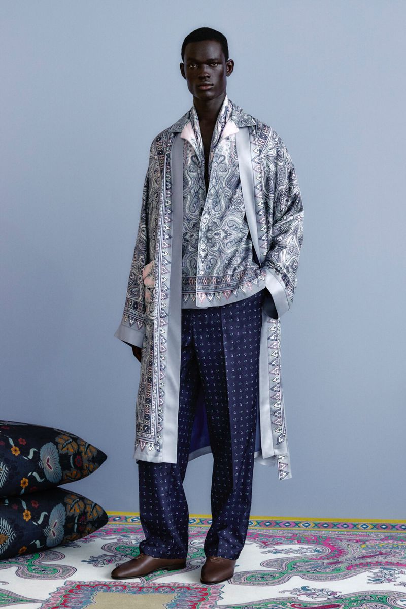 Man in paisley robe and patterned pants stands in front of a blue wall with pillows and a carpet featuring paisley patterns.