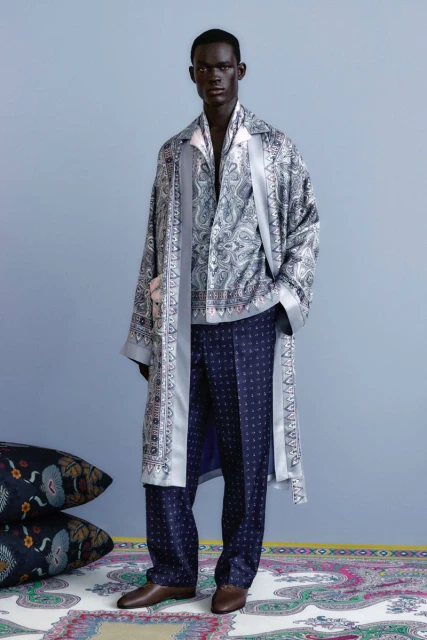 Man in paisley robe and patterned pants stands in front of a blue wall with pillows and a carpet featuring paisley patterns.