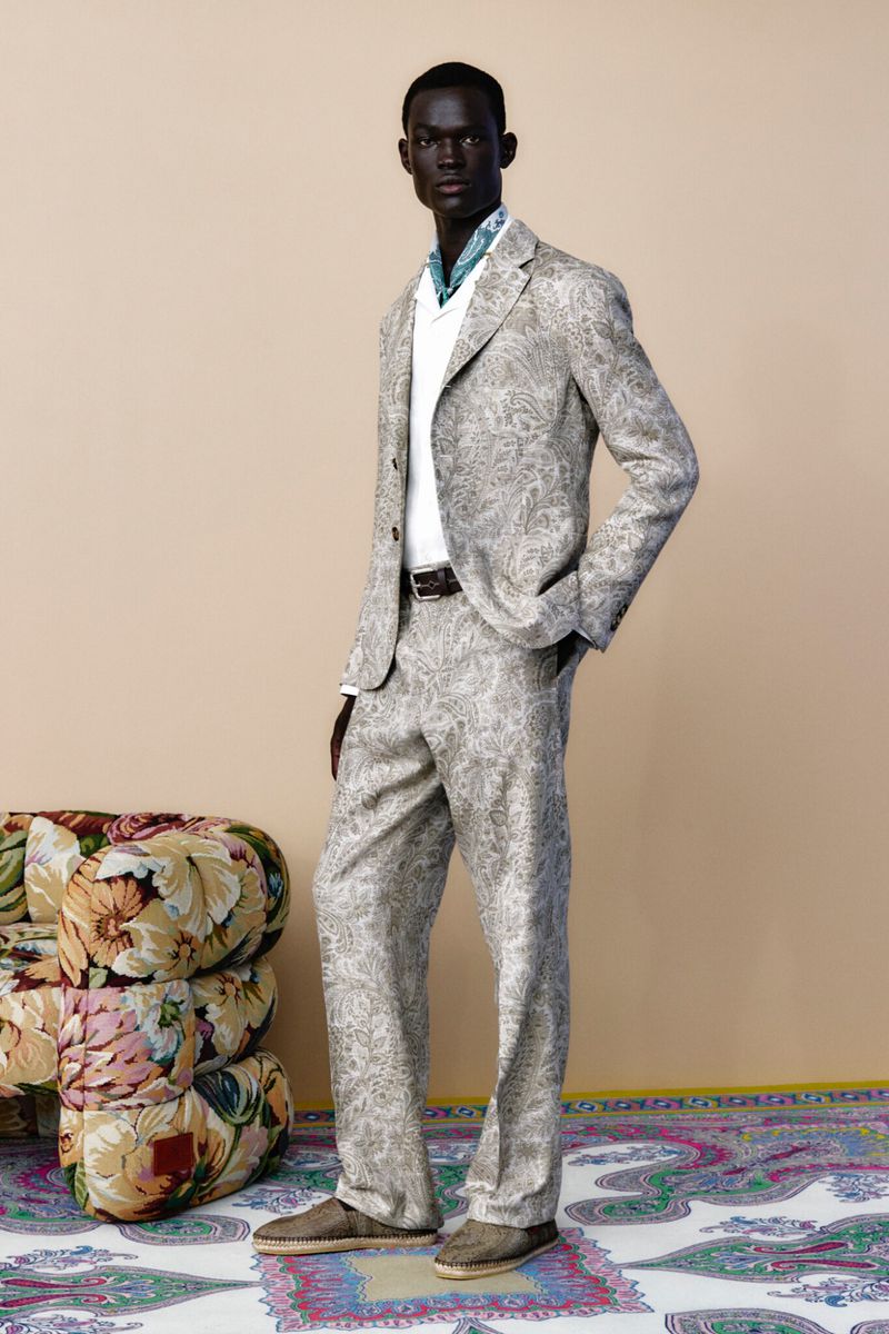 A dark-skinned male model stands in a paisley suit, a white shirt, and brown shoes against a neutral backdrop with a floral ottoman and rug.