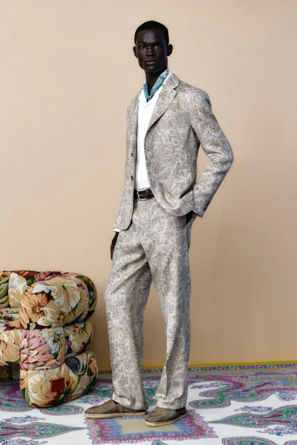 A dark-skinned male model stands in a paisley suit, a white shirt, and brown shoes against a neutral backdrop with a floral ottoman and rug.