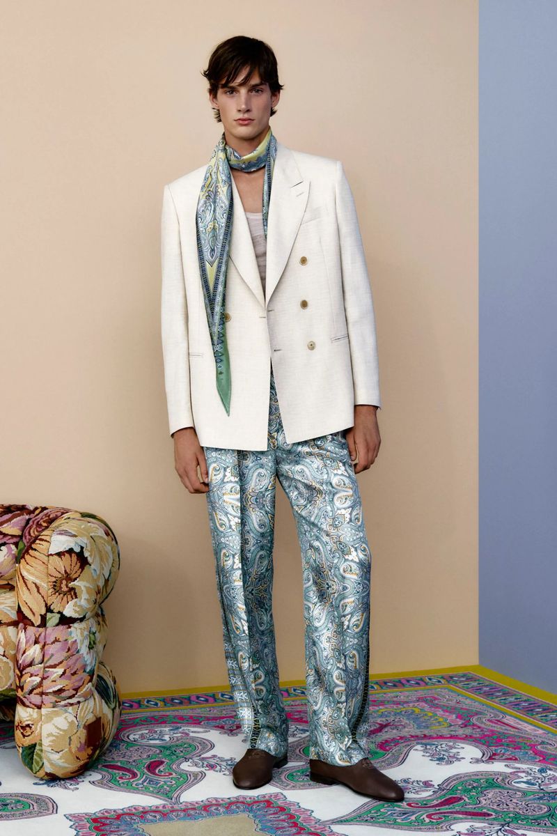 Adult male model poses in a cream-colored double-breasted suit with patterned pants and scarf, showcased against a pastel backdrop.