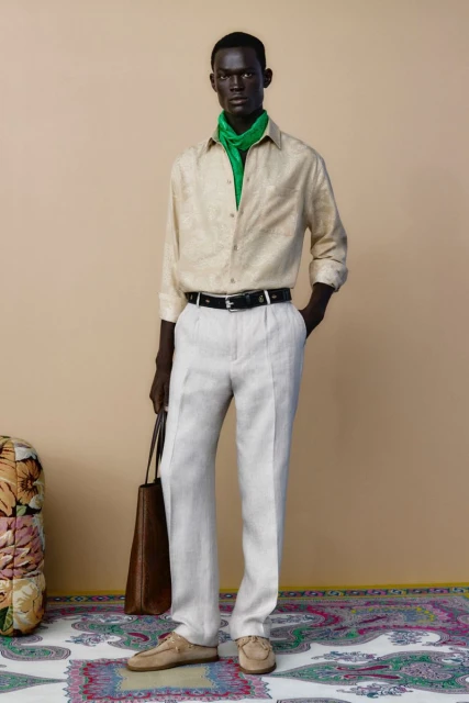 Full shot of an adult male fashion model in a button-down shirt, light trousers, and loafers, holding a bag and wearing a green scarf.