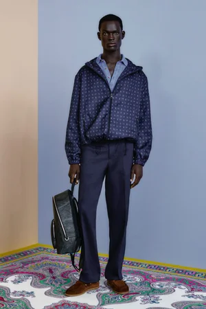 Model wearing a patterned dark blue jacket, blue shirt, dark trousers, holding a black backpack against a split-colour wall