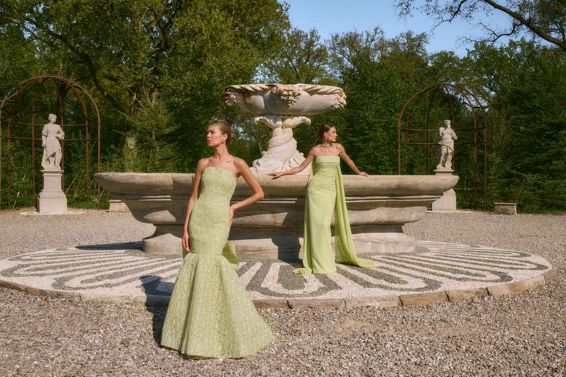 Two models wearing light green strapless evening gowns pose near a classical stone fountain in a formal garden courtyard.