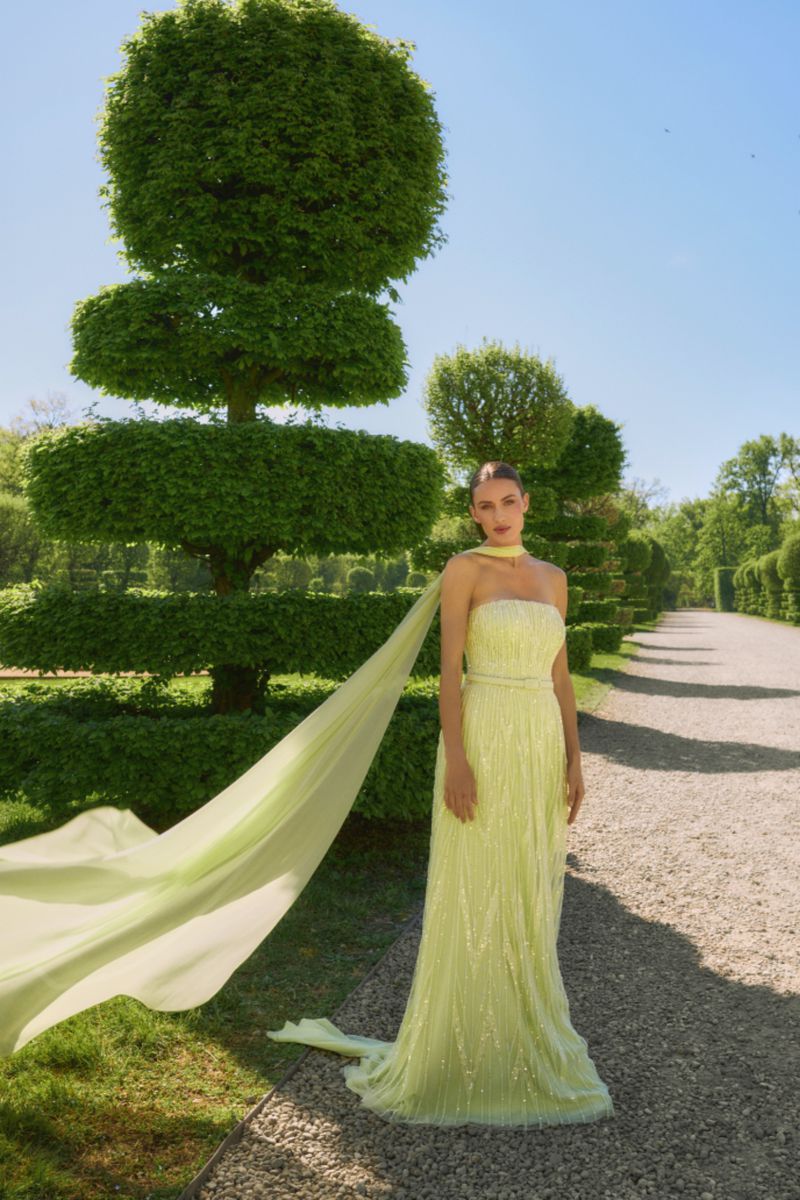 Model wearing a strapless pale yellow beaded gown with a flowing train along a gravel path beside sculptured topiary trees.