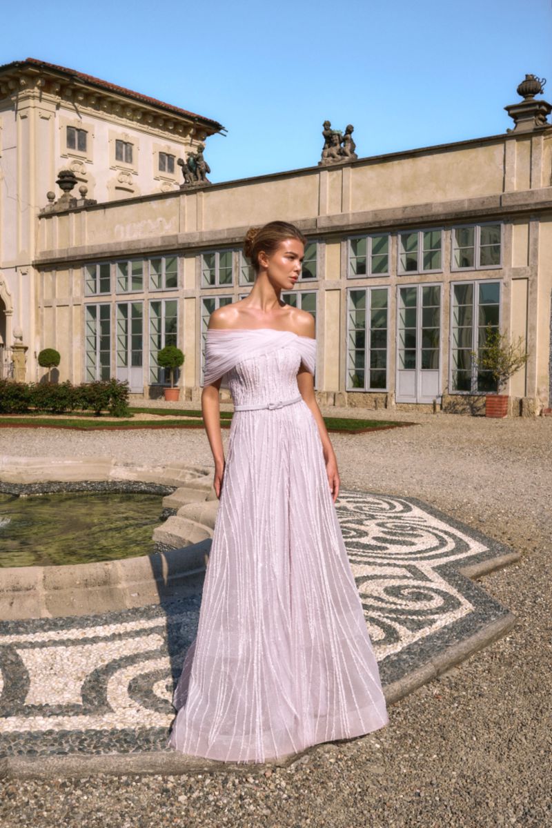 Model in off-the-shoulder lavender evening gown standing outdoors before a classical building and stone fountain.