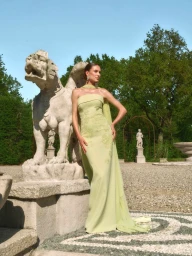 Model posing beside a carved stone beast statue wearing a strapless light green evening gown in an outdoor courtyard.