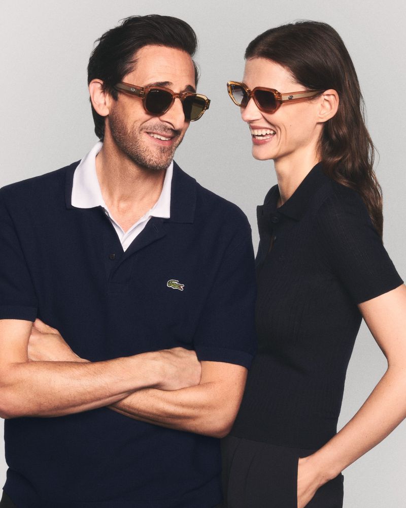 Adrien Brody and a smiling woman wearing tinted sunglasses and Lacoste polo shirts against a neutral studio backdrop.