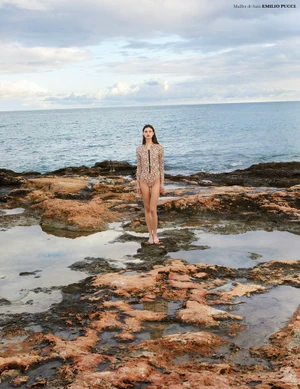 Model Sara Dijkink stands barefoot on a rocky shore wearing a patterned long-sleeved swimsuit by the sea.