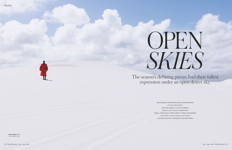 Fashion editorial spread showing a distant model in a vibrant red coat on expansive white sand dunes under a blue cloudy sky.