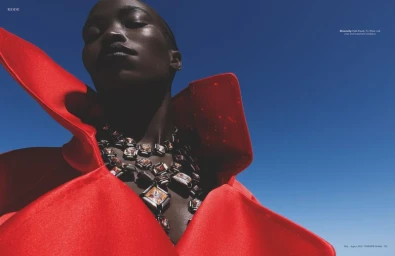 Low-angle shot of a Black woman wearing a sculptural red high-collar coat and ornate statement necklace against a clear blue sky