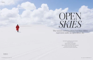 Fashion editorial spread showing a distant model in a vibrant red coat on expansive white sand dunes under a blue cloudy sky.