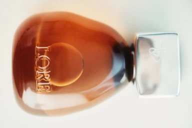 Amber glass Lore fragrance bottle with an organic pebble shape and polished silver cap on a white seamless background.