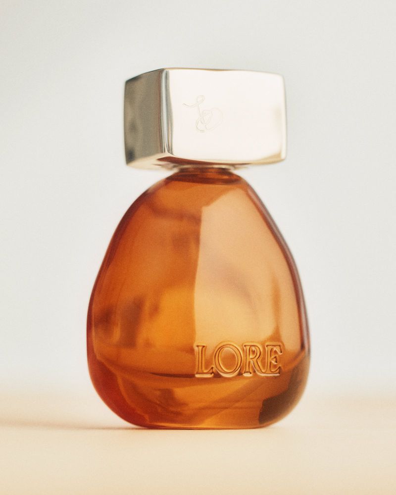 Sculptural amber glass perfume bottle with a metallic silver square cap and embossed Lore logo on a neutral surface.