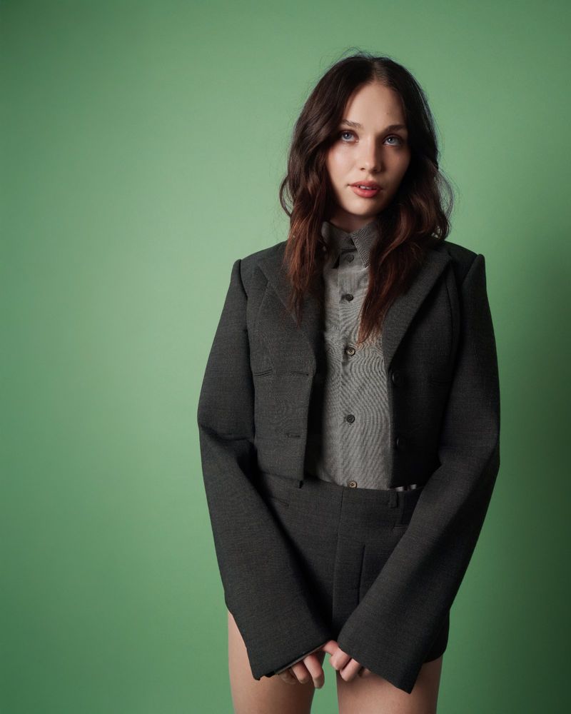 Maddie Ziegler posing in a charcoal cropped tailored blazer and shirt against a solid green seamless backdrop.