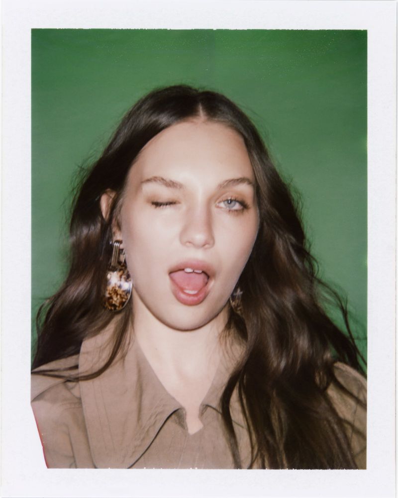 Maddie Ziegler winking and making an open-mouthed expression against a green backdrop in a collared shirt and statement drop earrings.