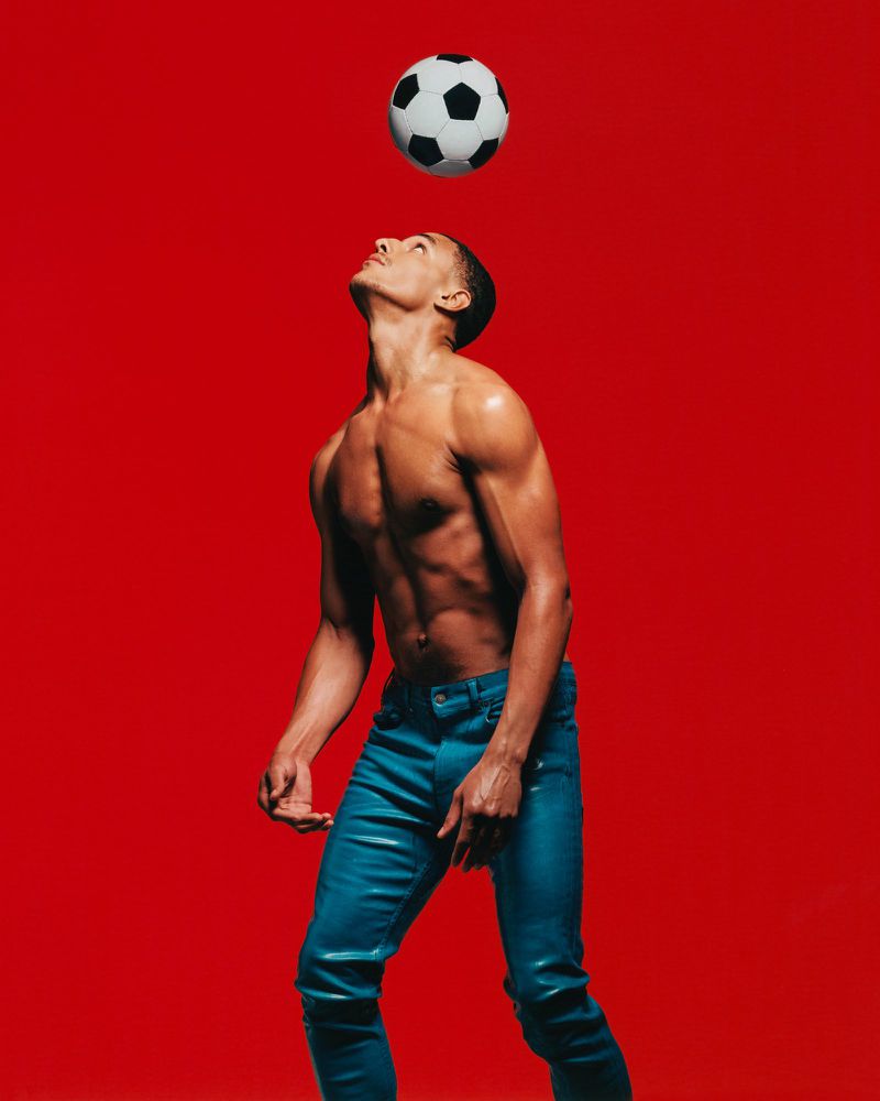 Shirtless male model wearing blue trousers balancing a soccer ball on his head against a bold red background.