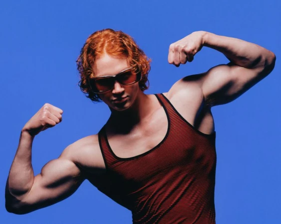 Muscular model with red hair wearing tinted sunglasses and a striped tank top flexing against a solid blue backdrop.