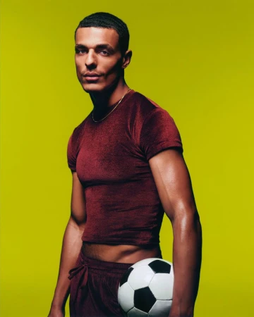 Model Taino Uppal in a maroon cropped t-shirt holding a soccer ball against a solid yellow-green studio background.