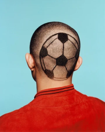Rear view of a man with short hair shaved and dyed into a graphic soccer ball pattern, wearing a red shirt against a cyan backdrop.