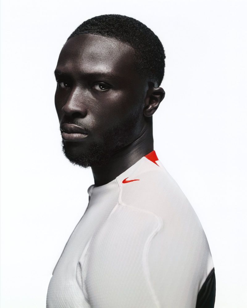 Portrait of a Black man looking at the camera wearing a white athletic top with red accents against a clean white seamless background.