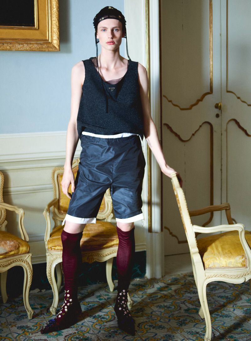 Model posing indoors in a dark sleeveless top, layered shorts, burgundy high socks, and a black skull cap near ornate yellow chairs.