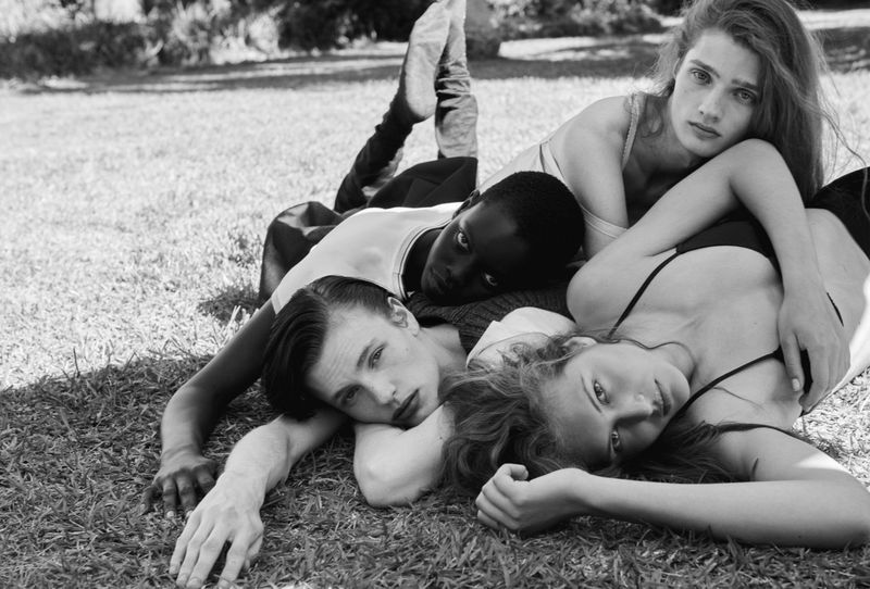 Black and white editorial photo of four models reclining together on a grassy lawn under bright natural daylight.