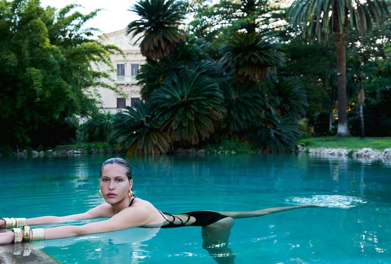 Model wearing a black cutout swimsuit and chunky gold cuffs swimming in an outdoor turquoise pool framed by palm trees.
