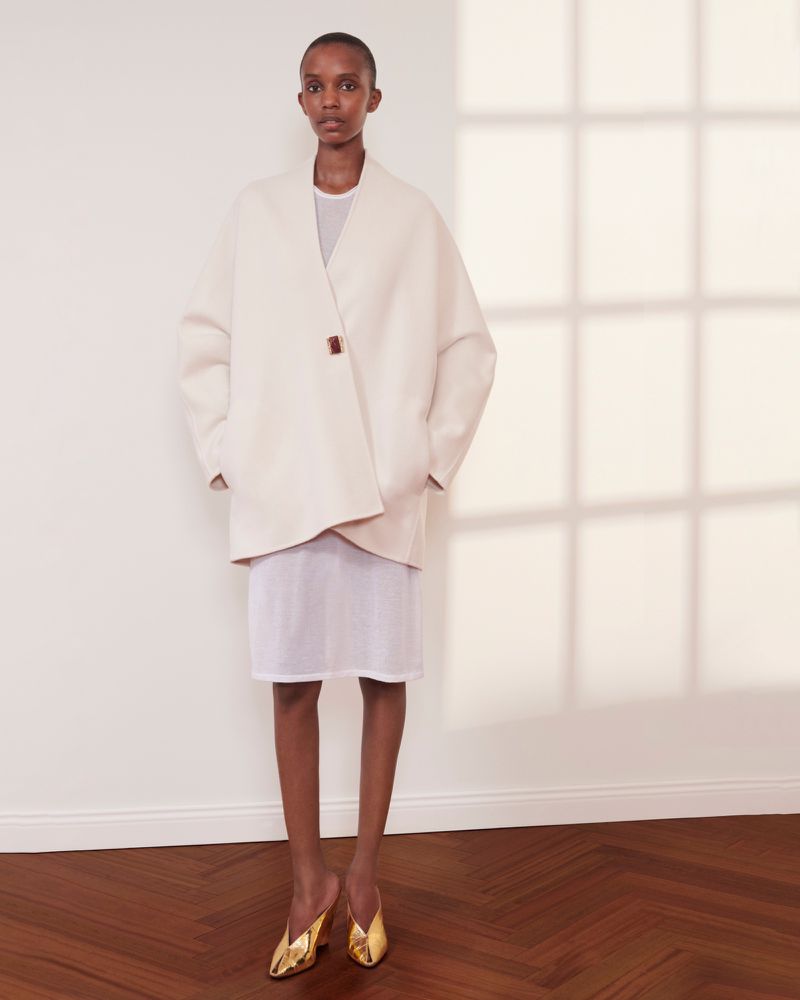 Elegant woman in a minimalist fashion shoot, wearing an off-white coat and gold shoes, standing against a plain white wall with shadow.