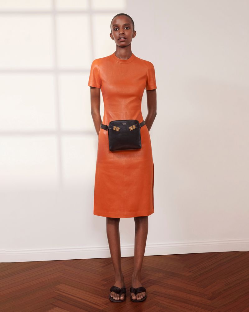 Indoor shot of a slim African-American woman posing in an orange dress with a black belt bag and black sandals against a white wall.