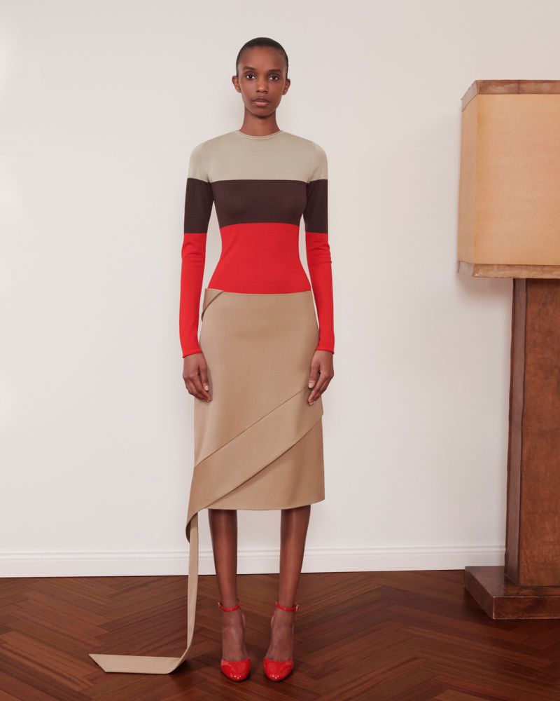 A woman poses in a color-blocked long-sleeve top and beige skirt with red heels, standing against a neutral background with wood floor.