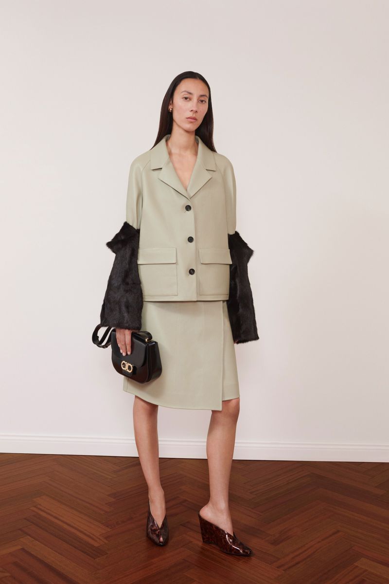 Fashion shot of a woman wearing a light green jacket and skirt, holding a black purse in a studio, standing against a white wall.