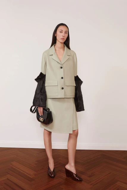 Fashion shot of a woman wearing a light green jacket and skirt, holding a black purse in a studio, standing against a white wall.