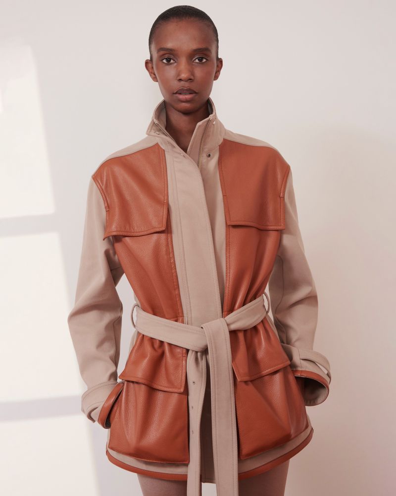 A Black female model poses in a light beige photo studio wearing a paneled tan and beige belted leather jacket, presenting a fashion editorial look.