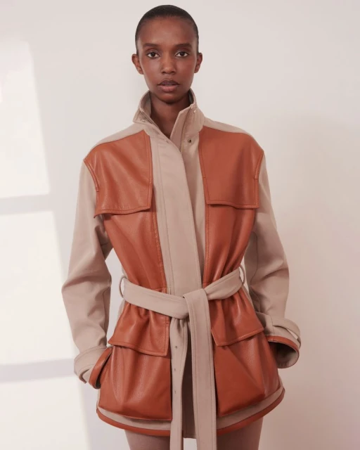 A Black female model poses in a light beige photo studio wearing a paneled tan and beige belted leather jacket, presenting a fashion editorial look.