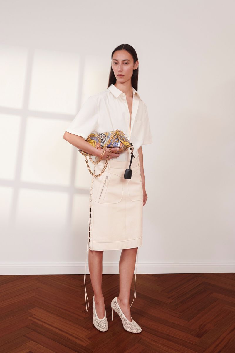 Model wearing a cream short-sleeved shirt, midi skirt, patterned clutch bag, and mesh heels against a white wall with parquet flooring.