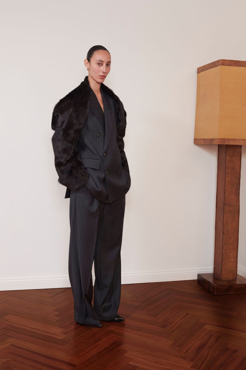 A woman in a pinstripe suit and fur jacket poses in an indoor setting with a brown herringbone floor and lamp.